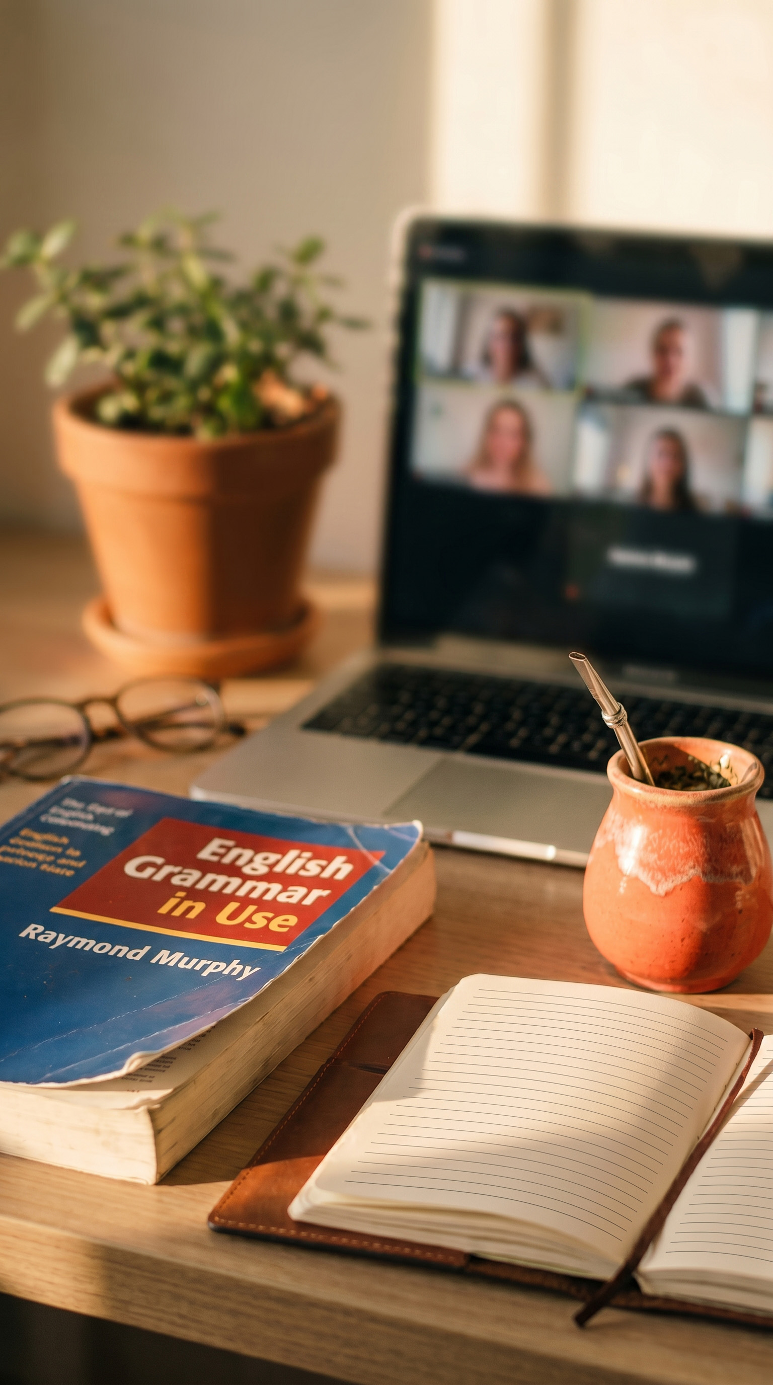 A teacher's desk during a live online English class — Murphy's English Grammar in Use, a notebook, mate gourd, and the class on a laptop